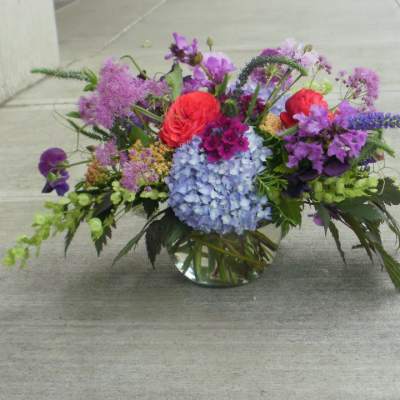 Low glass vase of mixed purple, blue, and red flowers arranged in a wide garden-style centerpiece