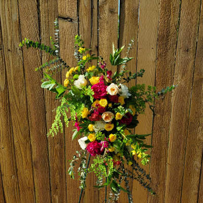 Bright standing spray of yellow, red, and white flowers on a metal easel against a wood fence.