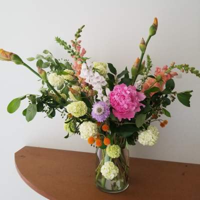 Tall mixed arrangement of pink peony, white hydrangea clusters, snapdragons, and orange accents in a clear glass vase