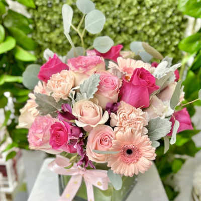 Pink and peach roses, carnations, and gerbera daisies in a glass cube vase with a pink ribbon bow