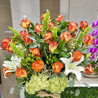 Tall arrangement of orange roses, white lilies, and green hydrangeas in a clear glass vase