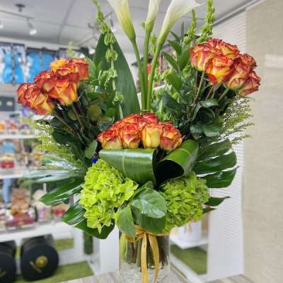 Tall arrangement of orange roses, white calla lilies, and green hydrangeas in a clear glass vase