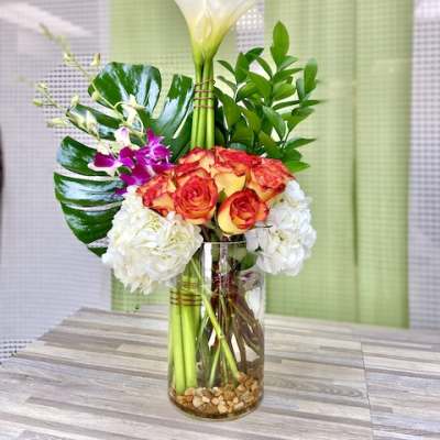 Tall arrangement of white calla lilies, orange roses, hydrangeas, and orchids in a glass vase with stones.