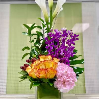 Tall modern arrangement with yellow roses, purple orchids, white calla lilies, and pink hydrangea in a glass cube vase