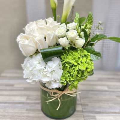 Tall white and green arrangement with calla lilies, roses, and hydrangeas in a glass cylinder vase