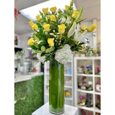 Tall arrangement of yellow roses, white lilies, and hydrangeas in a green-lined glass vase
