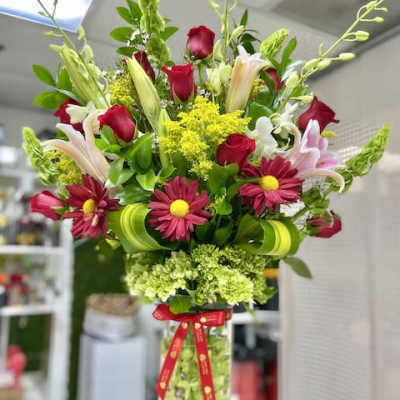 Tall mixed arrangement of red roses, pink lilies, and daisies in a clear cylinder vase with a red ribbon