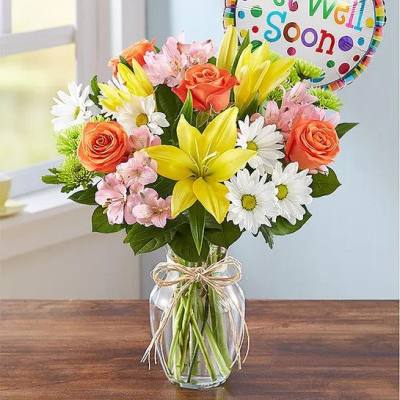 Mixed bouquet with yellow lilies, orange roses, white daisies and pink blooms in a clear vase with Get Well balloon