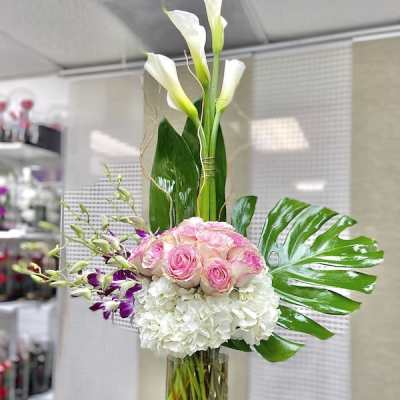 Tall arrangement of white calla lilies, pink roses, orchids, and hydrangeas in a clear cylinder vase.