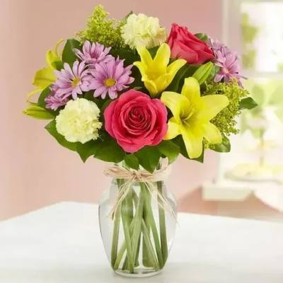 Mixed bouquet of pink roses, yellow lilies, purple daisies, and cream carnations in a clear glass vase