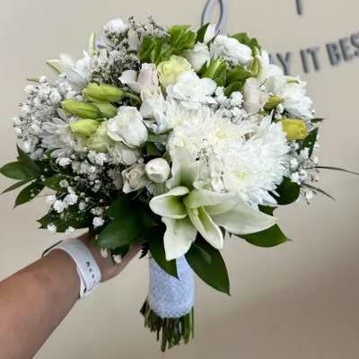 Hand-tied white bouquet with lilies, roses, and mums wrapped in white ribbon, held against a neutral background.