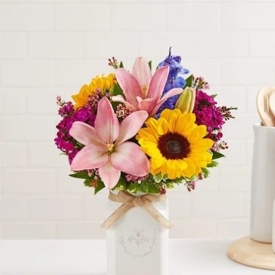 Colorful bouquet of pink lilies, a yellow sunflower, and mixed blooms in a white ceramic vase with a bow.