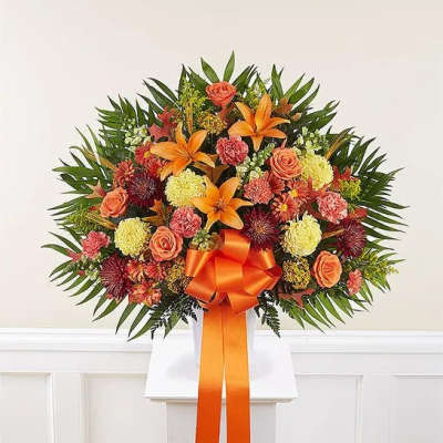 Fan-shaped arrangement of orange lilies, roses, carnations and mums with an orange bow in a white container