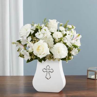 White flower arrangement in a white vase with a silver cross emblem on a wooden table