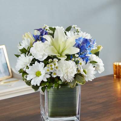 White lilies, daisies, and carnations with blue flowers in a square glass vase