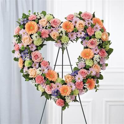 Heart-shaped standing wreath of pink roses, peach gerberas, and pastel daisies on a metal easel.