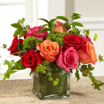 Low arrangement of red, pink, and orange roses with greens in a square glass vase