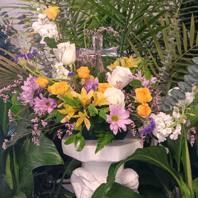 Arrangement of yellow and white flowers with a glass cross, set among tall palm greenery on a white pedestal.