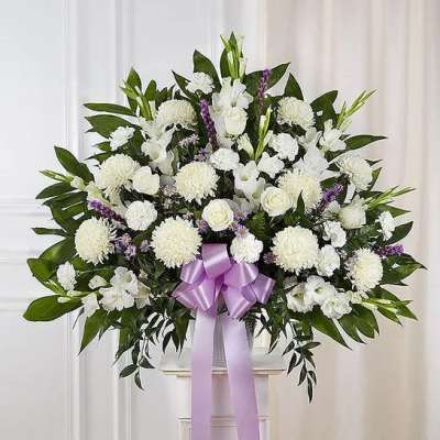 Large fan-shaped arrangement of white flowers with purple accents and a lavender bow on a white pedestal