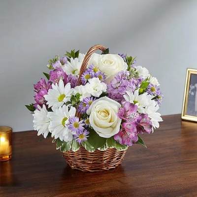 Basket of white roses, daisies, and purple flowers arranged in a small woven basket on a table.