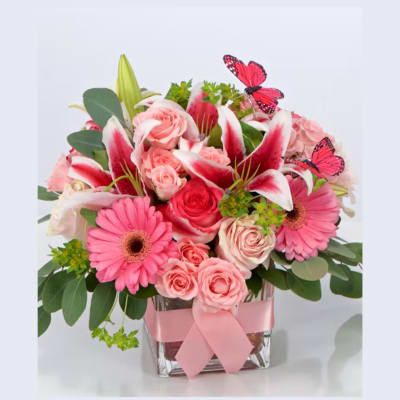 Pink lilies, roses, and gerbera daisies in a glass cube vase with pink ribbon and butterfly accents