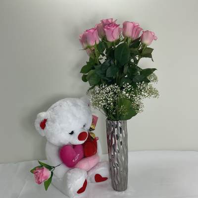 Pink roses in a tall silver vase beside a white teddy bear holding hearts