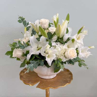 Low white and cream arrangement of roses, lilies, and hydrangeas in a white bowl on a gold pedestal stand