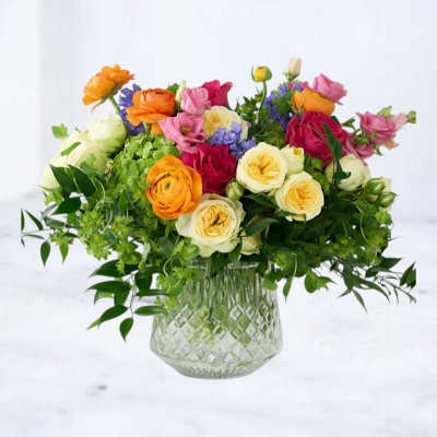 Colorful arrangement of yellow roses, orange ranunculus, and pink blooms in a clear patterned glass vase