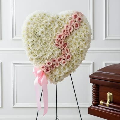 Heart-shaped white floral tribute with pink roses on a stand beside a casket