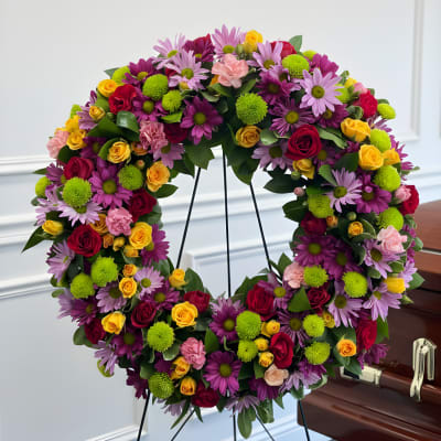 Large floral wreath with roses and daisies on a stand beside a casket