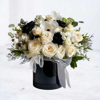 Round arrangement of ivory roses and white orchids with black blooms in a black hatbox with striped ribbon.