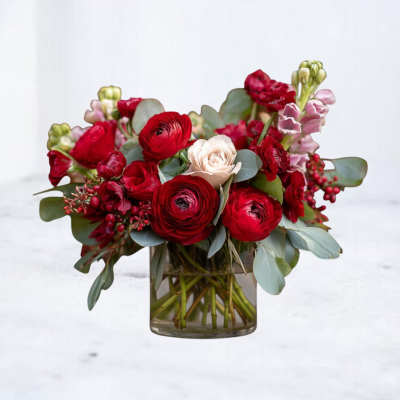 Low glass vase of red ranunculus and roses with a single pale bloom and pink accents.
