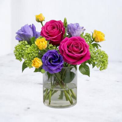 Short glass vase with hot pink roses, purple lisianthus, yellow blooms, and green hydrangea clusters.