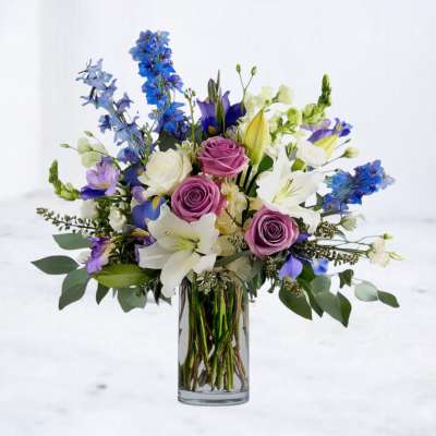 Tall glass vase of white lilies, purple roses, blue delphinium, and other mixed flowers.