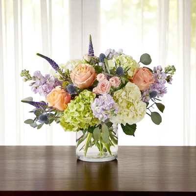 Peach roses, green and white hydrangeas, and lavender blooms in a clear glass cylinder vase