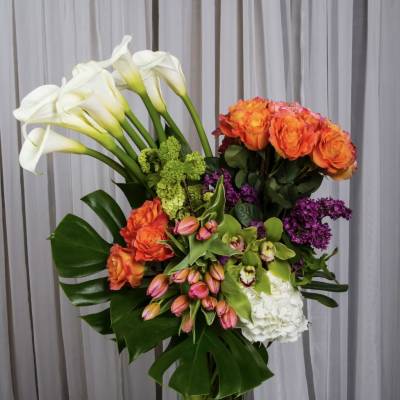Tall bouquet of white calla lilies, orange roses, and purple flowers in a glass vase