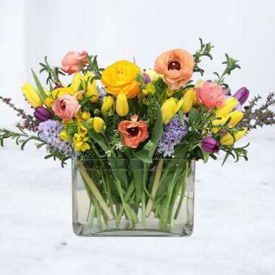 Low glass vase of yellow tulips, peach ranunculus, and purple hyacinths in a mixed spring arrangement