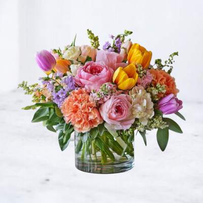 Low glass vase of pink peonies, tulips, orange carnations, and mixed pastel blooms.