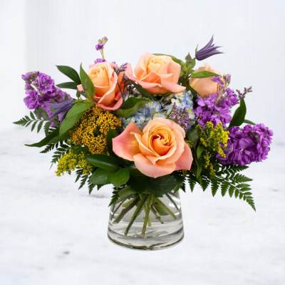 Short glass vase of peach roses with purple blooms, yellow accents, and blue hydrangea.