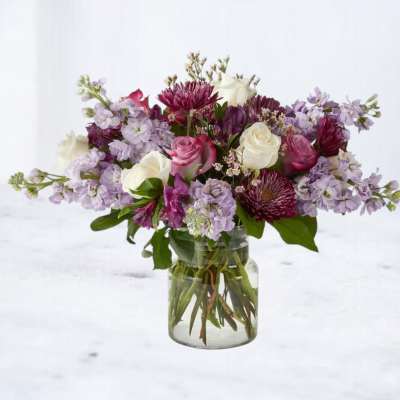 Mixed bouquet of purple, pink, and white flowers arranged in a clear glass jar vase