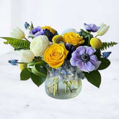 Low arrangement of yellow roses, blue hydrangeas, and purple flowers in a clear glass vase