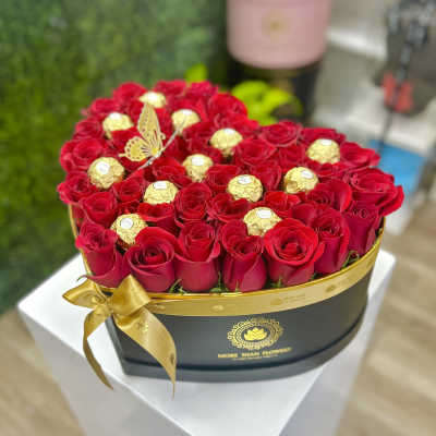 Heart-shaped black box of red roses with gold-wrapped chocolates and a gold ribbon bow.