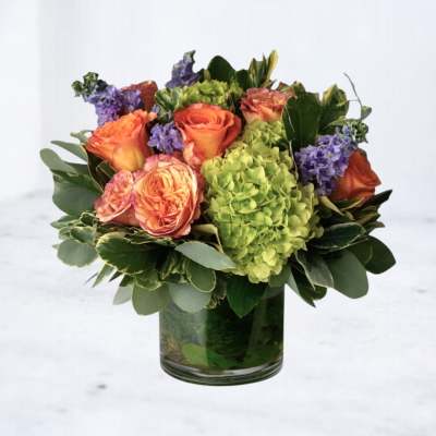 Low arrangement of orange roses, green hydrangeas and purple flowers in a clear glass vase