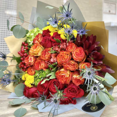 Hand-tied bouquet of red and orange roses with yellow blooms and blue thistles in kraft paper wrap