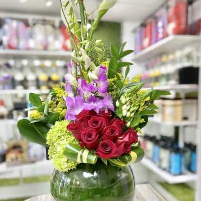 Tall arrangement with white calla lilies, purple orchids, and red roses in a round green-lined glass vase