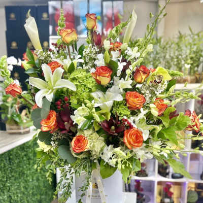 Tall hatbox arrangement with orange roses, white lilies, calla lilies, orchids, and green hydrangea
