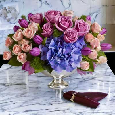 Pink roses and purple tulips arranged with blue hydrangea in a silver pedestal vase.