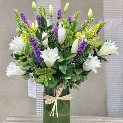 Tall arrangement of white lilies with purple and yellow flowers in a green glass vase