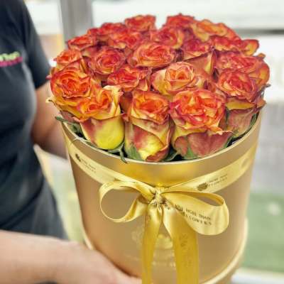 Round gold box of orange and yellow roses tied with a gold satin ribbon.