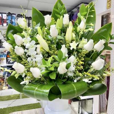 Large arrangement of white roses, lilies, and orchids in a white pedestal vase with bold green leaves.